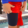 Boy by a pool wearing a blue rash guard and red NoNetz swim trunks showing a navy liner